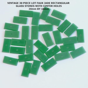 May include: A collection of thirty rectangular, faux jade glass stones with center holes. Each stone measures 20mm by 10mm. The stones are a vibrant green color and are scattered across a white surface. The text "VINTAGE 30 PIECE LOT FAUX JADE RECTANGULAR GLASS STONES WITH CENTER HOLES 20mm BY 10mm" is at the top.