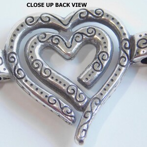 May include: Close-up back view of a silver-toned heart-shaped pendant with an intricate design. The pendant features a layered heart design with decorative swirls and dotted accents. Attached to the pendant are two silver chains.