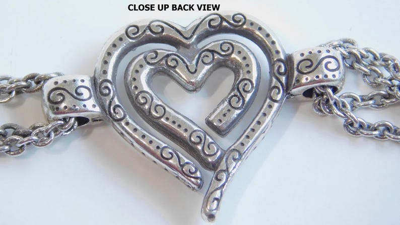 May include: Close-up back view of a silver-toned heart-shaped pendant with an intricate design. The pendant features a layered heart design with decorative swirls and dotted accents. Attached to the pendant are two silver chains.
