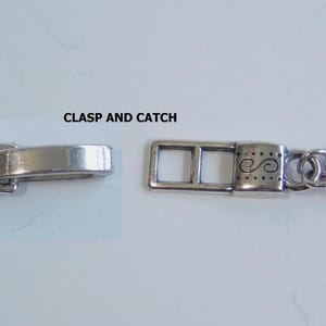 May include: Close-up of a silver-toned necklace clasp and catch. The clasp is rectangular with decorative swirls and dots. The catch is a simple, curved bar. The necklace chain is made of multiple strands.