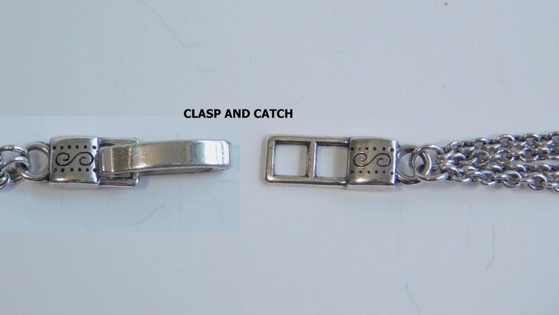 May include: Close-up of a silver-toned necklace clasp and catch. The clasp is rectangular with decorative swirls and dots. The catch is a simple, curved bar. The necklace chain is made of multiple strands.