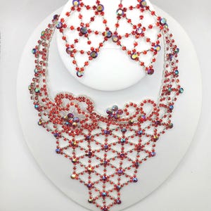 May include: A red rhinestone necklace and earrings set. The necklace is a delicate, intricate design with a v-shaped pendant. The earrings are dangling and feature a similar design.