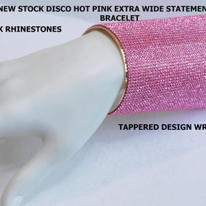 May include: A hot pink rhinestone cuff bracelet with a gold-tone band. The wide bracelet has a tapered design and is shown on a mannequin hand. The text on the image reads "VINTAGE NEW STOCK DISCO HOT PINK EXTRA WIDE STATEMENT GOLD TONE CUFF BRACELET".