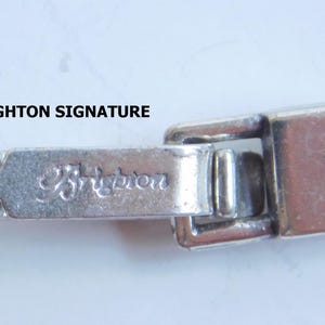 May include: Close-up of a silver-toned bracelet clasp with the engraved word "Brighton". The clasp is rectangular with a hinged closure and is connected to a chain of small, round links. The background is a light, neutral color.