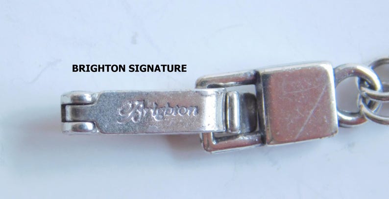 May include: Close-up of a silver-toned bracelet clasp with the engraved word "Brighton". The clasp is rectangular with a hinged closure and is connected to a chain of small, round links. The background is a light, neutral color.