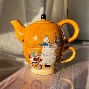Puede incluir: Juego de tetera y taza de cerámica en un tono naranja vibrante. El juego presenta un diseño caprichoso de un chef en bicicleta, llevando botellas de vino y una cesta. La tetera tiene un pomo negro en la tapa y un asa curva.