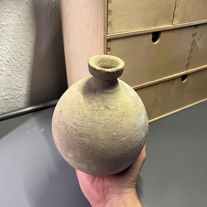 Puede incluir: Un pequeño jarrón redondo de cerámica de estilo antiguo, con un cuello estrecho y una amplia abertura circular. El jarrón es de color beige claro con una superficie rugosa y texturizada. El jarrón está sostenido en la mano de una persona.