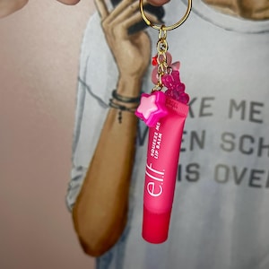 May include: A pink lip balm keyring with a gold-coloured ring. The keyring features a small tube of lip balm with the text "Squeeze Me Lip Balm" and the brand "elf." It is adorned with pink star and gummy bear charms.