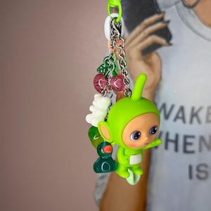 Può includere: Un portachiavi verde con un personaggio dei cartoni animati, un'anatra e ciondoli a forma di ciliegia. Il portachiavi ha un fermaglio verde e una catena argentata. Il personaggio ha occhi viola e una macchia bianca sul corpo.