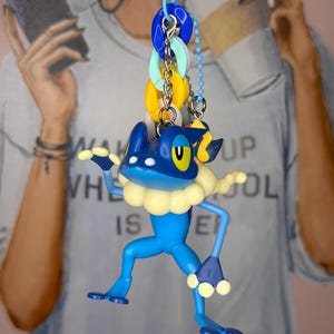 May include: A blue and yellow cartoon frog keychain with a light blue collar and yellow accents. The keychain is attached to a blue clasp with a series of colorful rings. The frog is suspended in front of a blurred background.