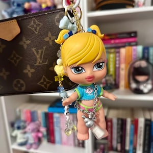 Puede incluir: Un llavero de muñeca con cabello amarillo, ojos azules, vistiendo un atuendo azul y verde, sosteniendo una botella plateada. La muñeca está unida a una cadena dorada con cuentas y dijes coloridos.