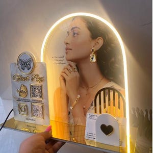 Può includere: Un espositore con l'immagine di una donna, illuminato da una luce calda. L'espositore presenta un logo a farfalla, icone di social media e il testo "Glam Box". La donna indossa gioielli d'oro, tra cui orecchini e un braccialetto.