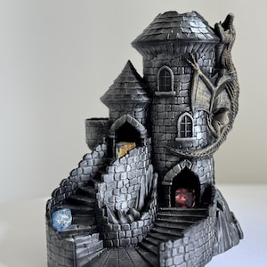 May include: A grey castle-shaped dice tower with a dragon sculpture. The tower features multiple levels, stairs, and arched windows. Dice of various colours are visible within the tower.