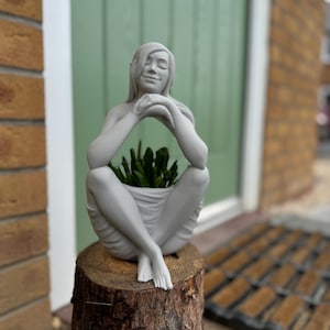 May include: A grey statue of a person in a seated pose, with arms crossed and legs crossed. The statue is a planter, holding green plants. It is placed on a tree stump, with a green door and brick wall in the background.