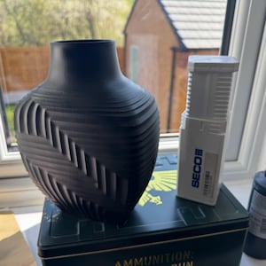 May include: A black vase with a textured pattern, alongside a white rectangular object and a black bottle. The items are displayed on a green metal box. The vase is a decorative item.