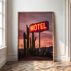 Pode incluir: Impress&atilde;o emoldurada com uma placa de motel vintage com a palavra "MOTEL" em n&eacute;on vermelho brilhante. A placa est&aacute; situada contra um c&eacute;u de p&ocirc;r do sol com cactos em primeiro plano. A imagem evoca uma paisagem des&eacute;rtica.