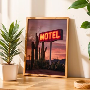 Pode incluir: Impress&atilde;o emoldurada com um letreiro de n&eacute;on "MOTEL" contra um p&ocirc;r do sol com cactos. A imagem est&aacute; em uma moldura de madeira, com uma planta em vaso &agrave; esquerda. O letreiro brilha com um tom laranja quente.