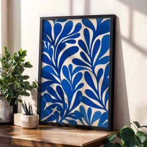 Op de afbeelding: Een ingelijste kunstdruk met een opvallend abstract ontwerp van blauwe, bladachtige vormen tegen een crèmekleurige achtergrond. Het kunstwerk is ingelijst in een zwart frame en wordt gepresenteerd op een houten oppervlak, met een potplant en potloden op de voorgrond.