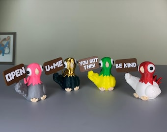 May include: A group of decorative pigeon figurines in various colours, each holding a sign with a different message. The pigeons are in pink, gold, green, and red, with signs that read "OPEN", "U+ME", "YOU GOT THIS!", and "BE KIND".