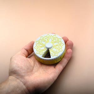 Pode incluir: Um organizador de comprimidos semanal branco e amarelo com um design floral na tampa. A caixa de comprimidos é redonda e tem compartimentos para cada dia da semana. Os dias da semana são impressos na lateral.