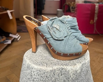 May include: A pair of denim high-heeled clogs with wooden soles and heels. The light blue denim uppers feature a flower embellishment and are accented with brown leather trim and silver studs. The heels are approximately 4 inches tall.