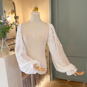 May include: A sheer white blouse with long, voluminous sleeves and delicate floral embroidery. The blouse is displayed on a mannequin with wooden hands, showcasing its design and fit. The sleeves have a ruffled cuff. The blouse is made of a lightweight fabric.