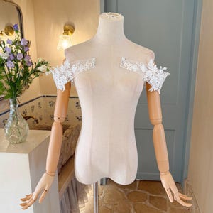 May include: A beige dress form with wooden arms and hands, displaying a delicate white lace shoulder piece. The lace features a floral design. The dress form is on a metal stand, set against a soft-toned background.