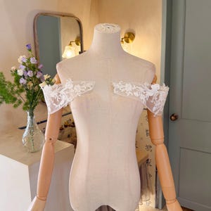 May include: A beige dress form with articulated wooden arms and hands. The form is adorned with white lace detailing across the shoulders. The background includes a mirror, flowers in a vase, and a light-colored wall.