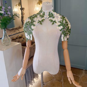 May include: A sheer, white lace shoulder wrap with intricate green leaves and white flower embroidery. The garment is displayed on a mannequin with wooden arms and hands, showcasing the delicate floral design. The high-collared wrap is fastened with small buttons.