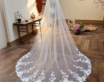 May include: A long, white bridal veil with a floral lace border. The veil is draped over a mannequin, showcasing its length and delicate design. The lace features detailed floral patterns.