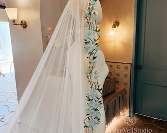 May include: A long, sheer white bridal veil draped over a mannequin, featuring an intricate vertical vine embroidery pattern. The design includes light blue and mint green fabric flowers, small white buds, and green stems along the edge.