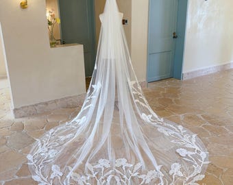 May include: A long, floor-length white bridal veil made of sheer tulle, featuring intricate white floral lace embroidery along the edges and the train. The delicate lace pattern depicts stems and blossoms that extend upward from the hem.