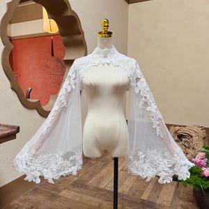 May include: A white lace bolero with long, sheer sleeves. The bolero features intricate floral embroidery along the edges and neckline. The garment is displayed on a mannequin.