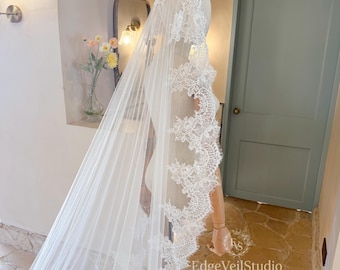 May include: A long, sheer white wedding veil draped over a mannequin. The veil features intricate, scalloped floral lace trim along the edges, extending from the head down to the floor. The delicate tulle fabric creates a soft, layered appearance.