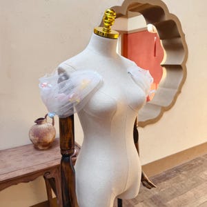 May include: A dress form with a white bodice and a gold-colored top. The form is adorned with a sheer, white, off-the-shoulder sleeve with colorful embellishments. The form is on a dark wooden stand.