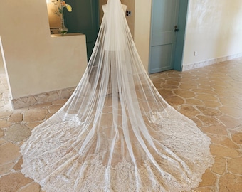 May include: A long, floor-length white bridal veil draped over a mannequin. The sheer tulle fabric features an intricate, wide lace border with detailed floral and scrollwork embroidery that extends along the entire hemline of the train.