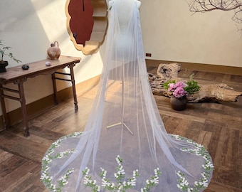 May include: A sheer white bridal cape with a floral embroidered hem. The cape is displayed on a mannequin, showcasing its flowing design. The hem features white flowers and green leaves.