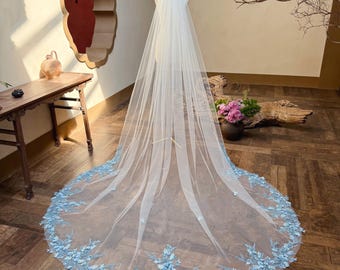 May include: A long, flowing white bridal veil with a delicate, light blue floral lace trim. The veil is displayed on a mannequin, showcasing its length and intricate design. The lace features detailed floral patterns.