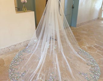 May include: A long, white bridal veil with delicate floral embroidery in shades of lavender, blue, and white. The veil is displayed on a mannequin, showcasing its flowing design and intricate details. The veil's train extends across the floor.