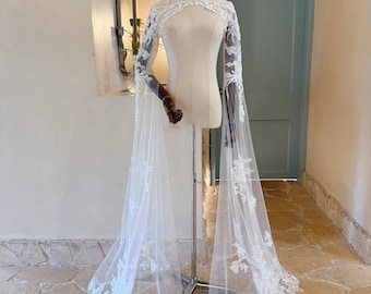 May include: A white bridal cape with long sleeves and a high neckline, displayed on a mannequin. The cape features intricate floral lace detailing along the sleeves and edges, with a flowing, sheer design. The text "EdgeVeilStudio" is visible.