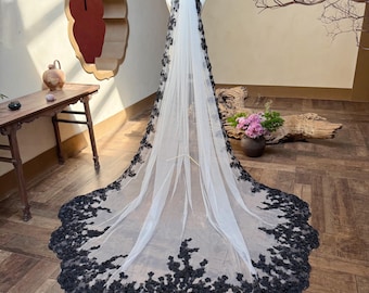 May include: A long, white bridal veil with a black floral lace trim. The veil is displayed on a mannequin, showcasing its length and intricate design. The black lace detailing adds a touch of elegance to the classic white veil.