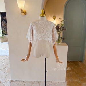 May include: A white lace bridal bolero with a delicate floral pattern and scalloped edges, displayed on a mannequin. The garment features a wide boat neckline and short, flowing sleeves that drape over the shoulders. Text reads EdgeVeilStudio.