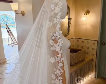 May include: A long, sheer white wedding veil draped over a mannequin, featuring intricate white floral lace embroidery that runs along the edge. The delicate lace pattern includes detailed blossoms and leaves, creating a continuous vine-like design.