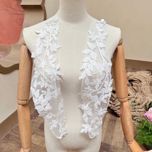 May include: Two white lace appliques with a delicate leaf and vine design. The appliques are displayed on a mannequin, showcasing the intricate floral pattern. The lace is a bright white, perfect for bridal or craft projects.