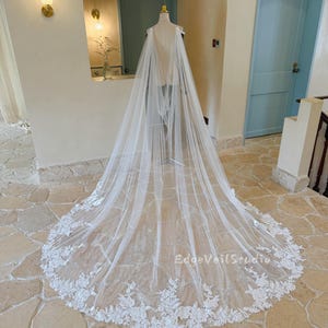 May include: A long, white bridal veil with a floral lace trim. The veil is displayed on a mannequin in a well-lit room. The text "EdgeVeilStudio" is visible on the veil.
