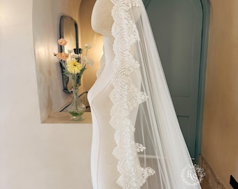 May include: A long, sheer white wedding veil draped over a mannequin, featuring an intricate scalloped lace border with detailed floral embroidery and delicate beadwork along the entire edge, extending down the length of the tulle fabric.