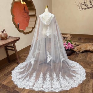 以下が含まれることがあります： 白いフード付きケープ、繊細なフローラルレースのディテール。 シアー生地のケープは、スカラップレースの縁取りが付いた長いトレーンが特徴です。 ケープはマネキンに展示されており、そのデザインと流れを強調しています。