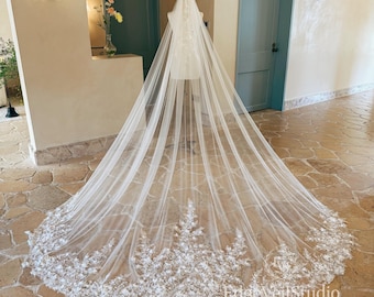 May include: A long, floor-length ivory bridal veil made of sheer tulle, featuring intricate white floral lace embroidery along the wide, scalloped hemline. The veil is draped over a mannequin, with the text EdgeVeilStudio visible at the bottom.