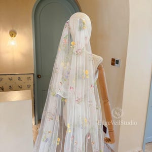 May include: A sheer white bridal veil featuring delicate, multi-colored floral embroidery in shades of yellow, pink, blue, and green. The veil is draped over a mannequin, showcasing the intricate botanical patterns scattered across the fabric.
