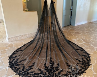 May include: A long black floor-length cape made of sheer tulle fabric, featuring intricate black floral lace embroidery along the wide, rounded hemline. The cape is draped over a mannequin, showcasing a deep V-neckline and a flowing silhouette.